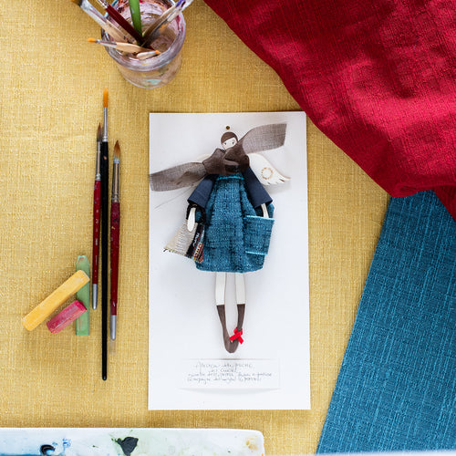 ANGELO delle AMICHE, struttura in legno dipinta e vestita a mano, abito in tessuto color blu con tascone, top in lana grigia e sciarpa in tessuto color marrone; borsetta sagomata in gobelin e piccolo fiocco di raso rosso sulla scarpetta. 
