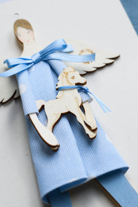 Dettaglio dell'ANGELO dei BIMBI, struttura in legno dipinta e vestita a mano, abito in cotone azzurro a pois e fiocco in raso; piccolo accessorio a forma di cavallo in legno con fiocco in raso azzurro.