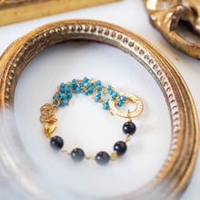 Load image into Gallery viewer, Dettaglio del bracciale ROSARIO realizzato per metà con un incatenato di perle di occhio di tigre blu e per l&#39;altra metà con una catena a rosario con cristalli turchesi; elemento centrale dorato a forma di cerchio.
