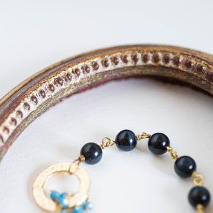 Dettaglio del bracciale ROSARIO realizzato per metà con un incatenato di perle di occhio di tigre blu e per l'altra metà con una catena a rosario con cristalli turchesi; elemento centrale dorato a forma di cerchio.