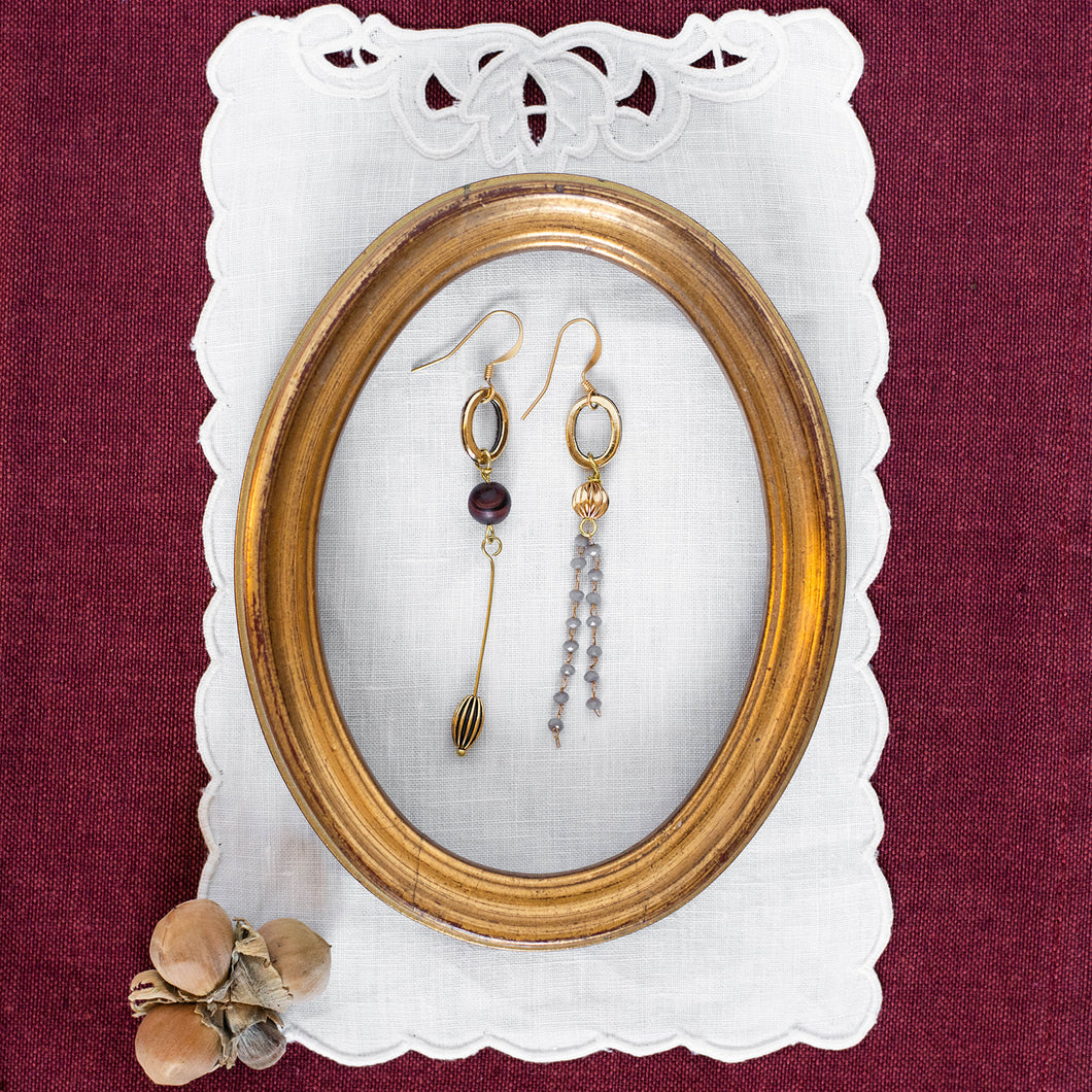 Orecchini FILI asimmetrici con due ovali dorati, perle di occhio di tigre rosso satinato e charms dorati; catena pendente a rosario con cristalli grigi.