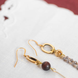 Dettaglio degli orecchini FILI asimmetrici con due ovali dorati, perle di occhio di tigre rosso satinato e charms dorati; catena pendente a rosario con cristalli grigi.