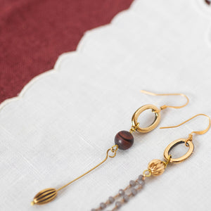 Dettaglio degli orecchini FILI asimmetrici con due ovali dorati, perle di occhio di tigre rosso satinato e charms dorati; catena pendente a rosario con cristalli grigi.