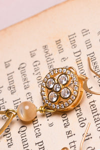 Dettaglio degli orecchini QUADRIFOGLIO asimmetrici con un elemento metallico tondo con struttura interna a forma di quadrifoglio con zirconi sfaccettati ed incastonati, goccia pendente e perlina in madreperla cappuccino.